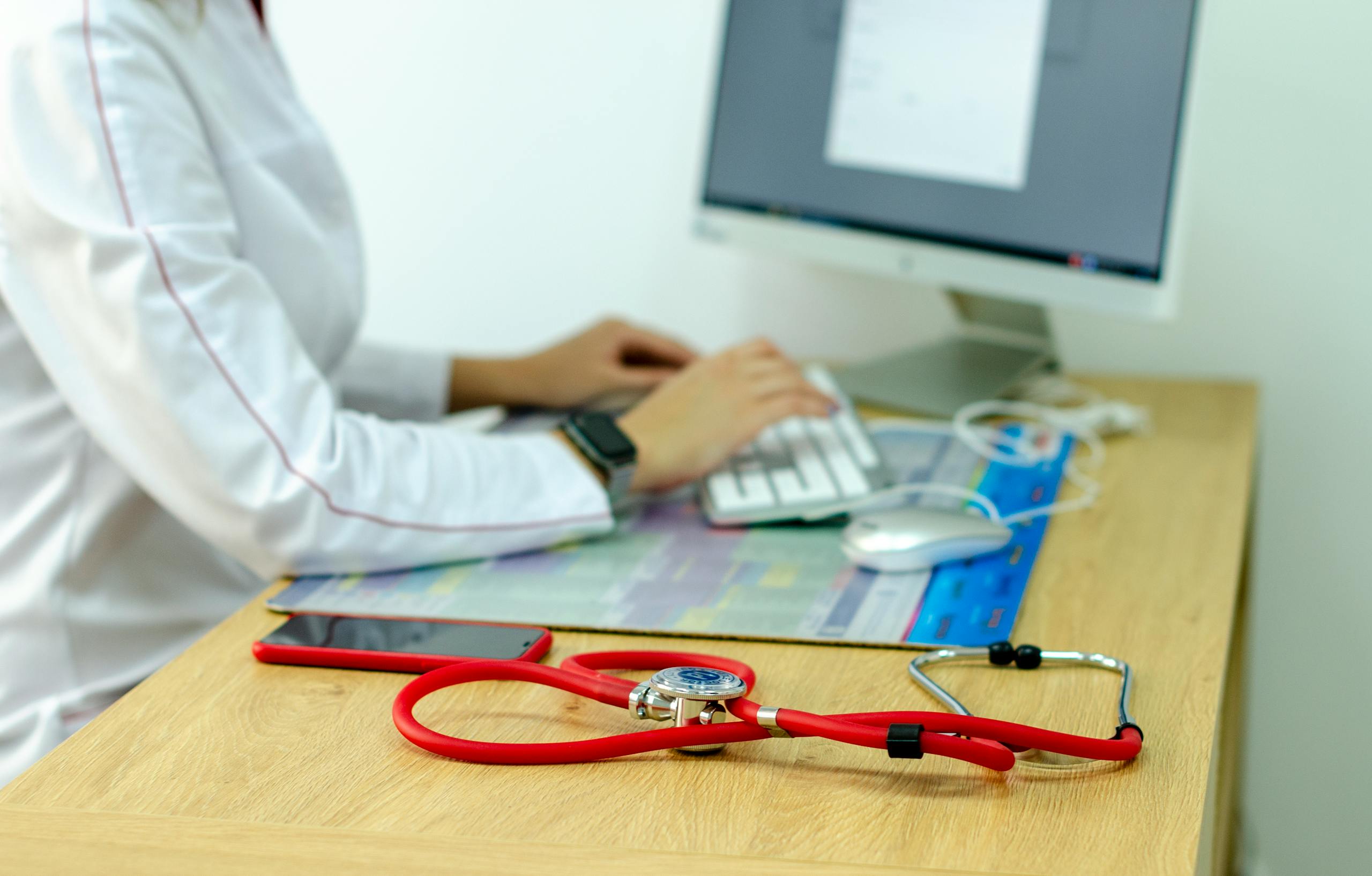 A medical professional typing on a computer with a stethoscope on a wooden desk, indicating a modern healthcare setting.